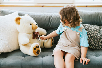 cute little curly haired girl plays doctor with a stethoscope and treats and cares a teddy bear on a sofa