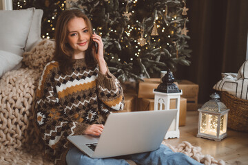 Young beautiful woman with red hair in cozy sweater using mobile smartphone video call to family and friends in room with christmas tree at home