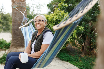 Beautiful senior blonde woman reading book and sitting in hammock in the garden