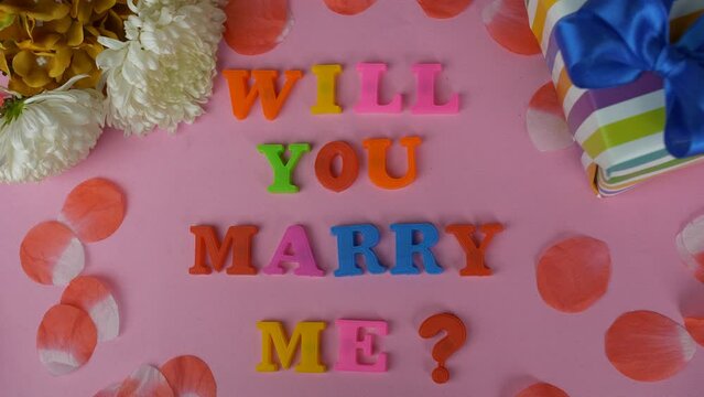 Will You Marry Me Marriage Proposal- decorated with flowers  word formation. A colorful gift box with a "Will You Marry Me" phrase against a pink background - propose 