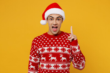 Merry proactive young man wear knitted christmas sweater Santa hat posing hold index finger up with...