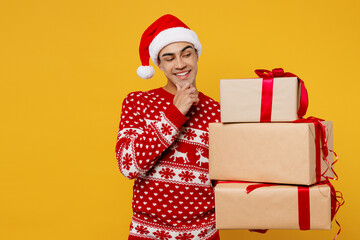 Merry minded young man wear knitted christmas sweater Santa hat posing hold many present box with gift ribbon bow isolated on plain yellow background. Happy New Year 2023 celebration holiday concept.