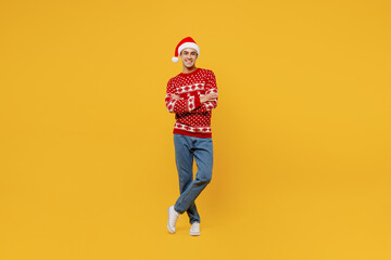 Fototapeta na wymiar Full body merry young man wear red knitted Christmas sweater Santa hat posing hold hands crossed folded look camera isolated on plain yellow background Happy New Year 2023 celebration holiday concept