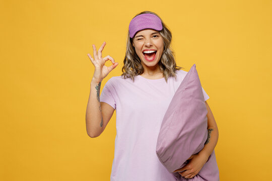 Calm young woman she wears purple pyjamas jam sleep eye mask rest relax at home show ok okay gesture hold pillow wink isolated on plain yellow background studio portrait. Good mood night nap concept.