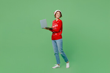 Full body merry young IT woman wear knitted xmas sweater Santa hat posing hold use work on laptop pc computer isolated on plain pastel light green background. Happy New Year 2023 celebration concept.