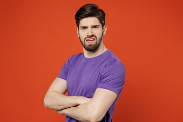 Fototapeta premium Side view young brunet caucasian man 30s in casual basic purple t-shirt look camera hold hands crossed folded look camera isolated on plain orange background studio portrait. People lifestyle concept.
