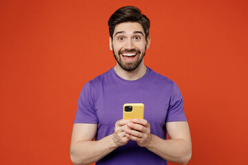 Young surprised brunet caucasian man wear casual basic purple t-shirt hold in hand use mobile cell phone in yellow case isolated on plain orange background studio portrait. People lifestyle concept.