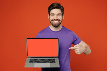 Young brunet cheerful IT man 30s wear casual basic purple t-shirt hold use work point finger on laptop pc computer with blank screen workspace area isolated on plain orange background studio portrait