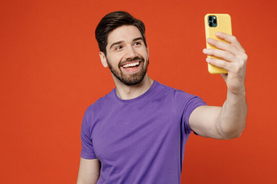 Young brunet man 30s wear casual basic purple t-shirt doing selfie shot on mobile cell phone post photo on social network isolated on plain orange background studio portrait. People lifestyle concept.