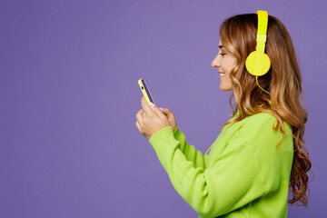 Side view young woman 30s wear casual green knitted sweater headphones listen to music dance gesticulating hands have fun isolated on plain pastel purple background studio. People lifestyle concept