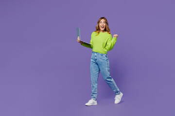 Full body young fun side view IT woman 30s she wears casual green knitted sweater hold use work on laptop pc computer walk do winner gesture isolated on plain pastel purple background studio portrait.