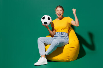 Fototapeta na wymiar Full body young fun woman fan wear basic yellow t-shirt cheer up support football sport team sit in bag chair hold soccer ball watch tv live stream do winner gesture isolated on dark green background.