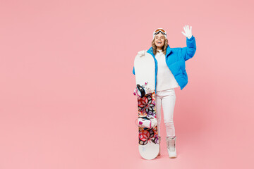 Full body snowboarder woman wear blue suit goggles mask hat ski padded jacket hold snowboard waving hand isolated on plain pastel pink background Winter extreme sport hobby weekend trip relax concept