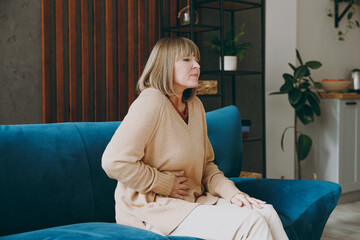 Elderly woman 50s years old wears casual clothes sit on blue sofa put hand on abdomen suffer from...