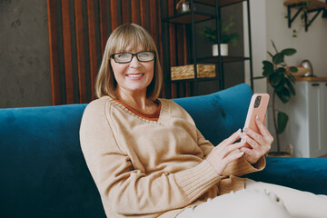 Obraz premium SIde view elderly happy woman 50s years old wear casual clothes sits on blue sofa hold in hand use mobile cell phone stay at home flat rest relax spend free spare time in living room indoor grey wall
