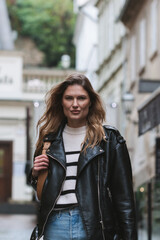 Young street fashion blond woman walking toward in urban city environment, serious face, determined,  looking at camera, black leather jacket and blue jeans. Waist up portrait crop.