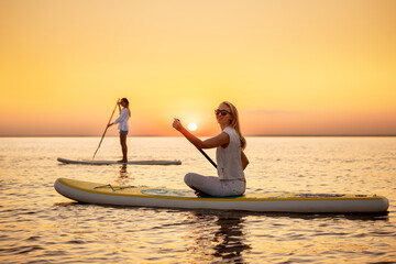 Two happy slim girls are walking on sup stand up paddle boards at sunset calm lake