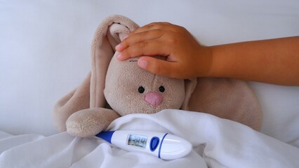 Child's little hand touches forehead of a sick toy bunny hare lying in bed with a high fever to check temperature with a thermometer. Medicine, healthcare and childhood concept