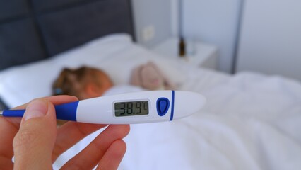 Close-up thermometer. Mother measuring temperature of her ill kid. Sick child with high fever laying in bed and mother holding thermometer.
