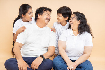 Happy indian grandparents with kids wearing white casual t-shirt sitting together looking at each other and laughing having a good time isolated beige background.