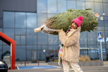 Woman in winter clothes carries wrapped Christmas tree on shoulder near shopping mall outdoors....