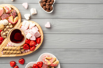 Valentine's Day concept. Top view photo of wooden heart shaped serving tray glass cup of drinking saucers with sweets chocolate jelly candies and cookies on grey wooden desk background with copyspace