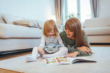 Smiling young mother sit on warm floor play with little infant toddler child, happy biracial mom relax have fun read book with small baby girl at home, motherhood, childcare concept