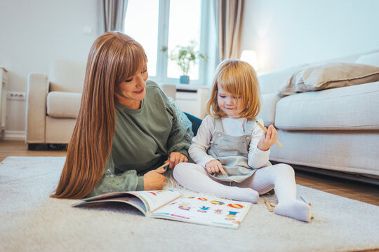Shot of a young beautiful mother reading a book to her adorable daughter in bed at home. Mother and daughter lying on warm floor reading book. Little girl working her homework with mother.