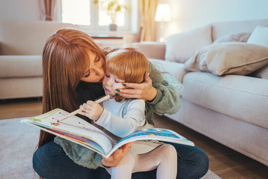 Attractive mother with little adorable daughter lying together at cushions on wooden warm floor in living room at home reading a book fairy tale close up. Weekends activities and development concept