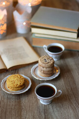Plates with various cookies, cups of tea or coffee, vintage books, reading glasses and lit candles on the table. Selective focus.
