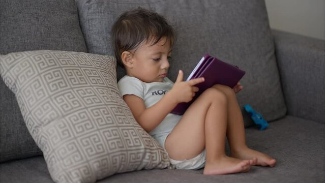 Young little latin baby boy in a light blue baby romper sitting on a grey couch enjoying a movie on a purple tablet