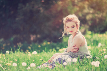Fototapeta na wymiar Beautiful girl in a vintage look sits at sunset in a field of dandelions and holds a hat in her hands, spring
