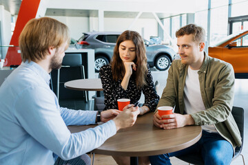 A car dealership manager advises a young couple before buying a new family car at a dealership. A man and a young woman drink coffee and listen to the manager