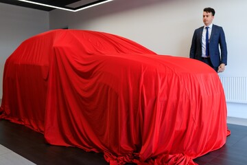 The car is covered with a red cloth before sale. In the background is the sales manager of the dealership