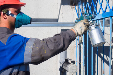Industrial painter in respirator and helmet paints metal fence from spray gun. Painting fence on summer day. Authentic workflow. Professional worker in overalls..