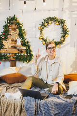 A young woman in glasses sits on a bed in a home environment and works remotely. New Year. New Year's decor in the room