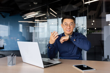 Asian businessman uses smartwatch to communicate, man gives commands to artificial intelligence using wrist watch, boss in shirt works inside office using laptop. - Powered by Adobe