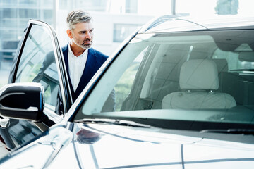 Male driver opening door while look the interior of his new auto. Seriously beard grey hair man finally gets long-awaited car, wearing formal suit.