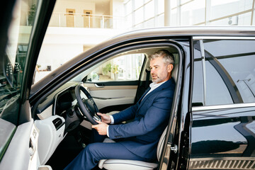 Fototapeta na wymiar Side view of cheerful handsome senior senior man in formalwear sitting in car and looking the interior of his new auto in dealership..