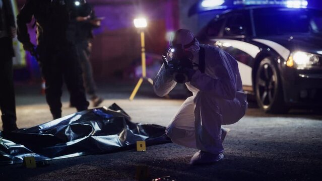 Man from Forensics Team Taking Photos of Evidence and Victim's Body in a Crime Scene at Night. Professional Investigators and Police Officers Working to Solve the Case of Potential Premeditated Murder