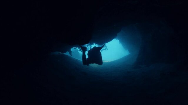 Diver swimming in underwater cave tunnel, back view. Scuba diving in ocean, undersea marine life exploring, sunlight shining through dark cavern