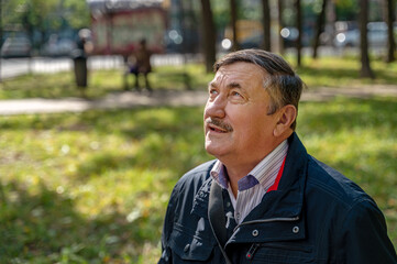 Portrait of a pensive senior man sitting on the bench, in the public park, outdoors. Old man relaxing outdoors and looking away. Portrait of elderly man enjoying retirement