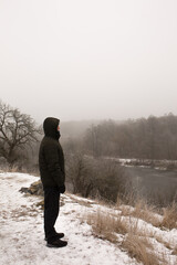 A man stands on a high cliff near a river in winter