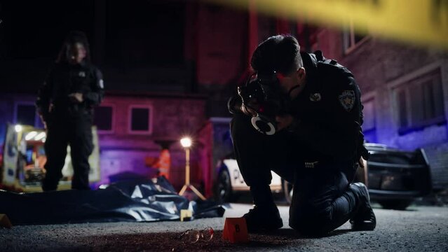 Asian Policeman Taking Pictures of Marked Evidence While Police Female Chief and Detective Talk about the Victim's Bagged Corpse in the Background. Bloody Glasses and Murder Weapon on the Floor