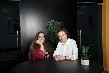 Girl talking her best friend. Two women looking at camera sitting at table in office against dark backdrop. Beautiful smiling brunette big size woman with brown long hair. Love of Best Friends.
