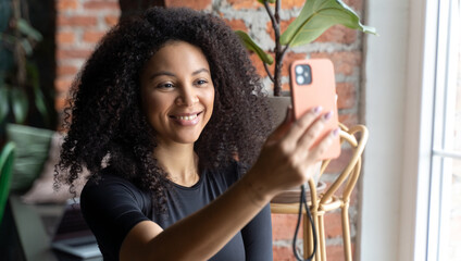 Black happy woman with African curls speaking video chat, call on smartphone at home or cafe indoors. Communication with friends of relatives. Comfort relationships. Freelance