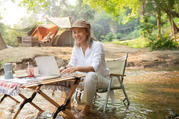 Fototapeta premium Asian woman travel and camping alone reading a book and using laptop while relaxing on the deck chairs in the river at natural park in Thailand. Recreation and journey outdoor activity lifestyle.