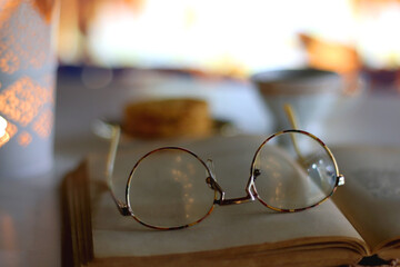 Decorative candle holder, cup of tea or coffee, plate of cookies, open book and reading glasses in front of the fireplace. Selective focus.