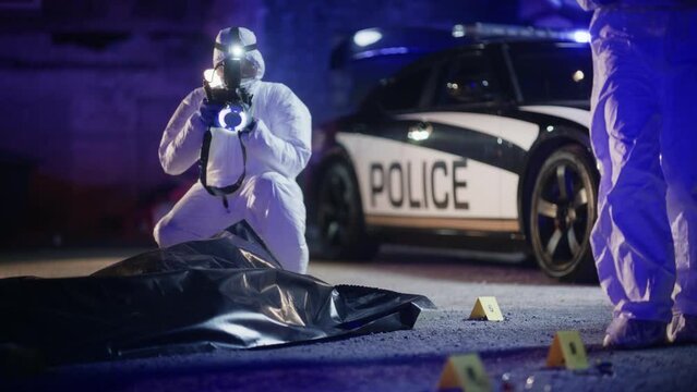 Portrait of Two Forensics Experts Doing Fieldwork at Night at a Crime Scene. One Technician Taking Photos of the Dead Body While the Other Packs the Bloodied Knife as Murder Weapon.