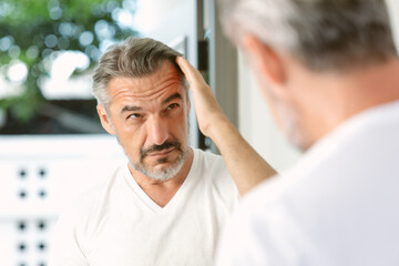 Handsome middle age caucasian man with beard looks in the mirror. Middle-aged man concerned by hair loss. Men's personal care. Morning routine. Receding hairline, haircare, beauty concept